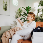 A woman sitting on a couch surrounded by clothes, appearing thoughtful and overwhelmed, reflecting the long-term emotional impact of growing up with narcissistic parents.